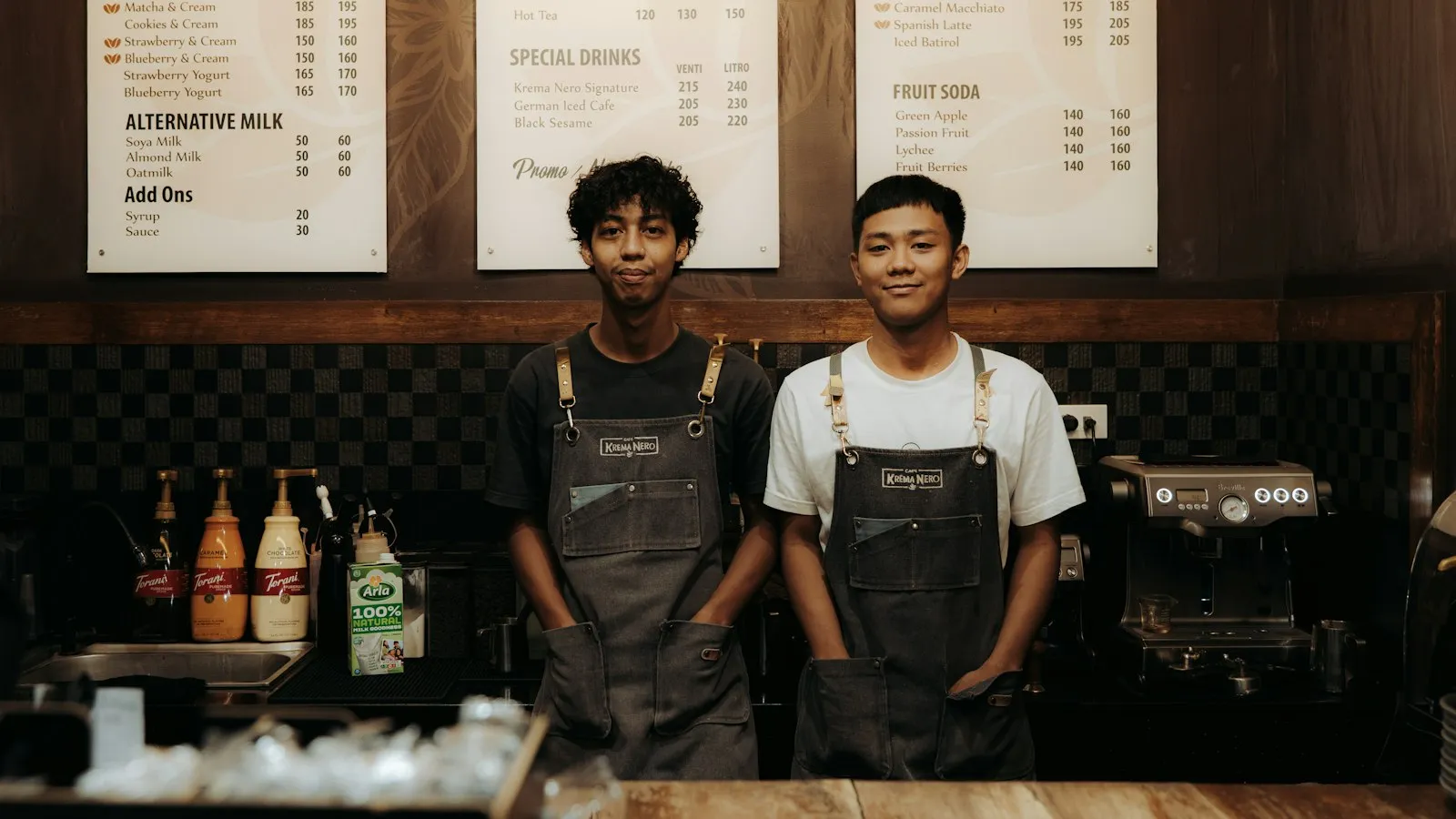 Two baristas in aprons standing behind a cafe counter at shift change